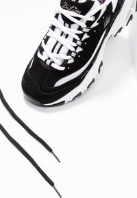 Black and white athletic shoe featuring a textured upper, white laces, and a robust sole with black accents. Two black shoelaces are included.
