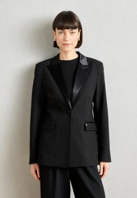Černý blazer s saténovým klopovým límcem, dvěma předními kapsami, zapínáním na jeden knoflík a přiléhavým střihem, v kombinaci s černým topem.