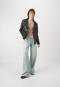 Giacca da biker in pelle nera, pantaloni in denim azzurro chiaro larghi, top rosa a scollo a V. Capelli ricci, in posa eretta, sfondo minimalista.