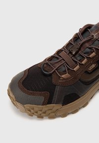 Brun atletisk sko med teksturert stoff og gummisåle. Har elastiske snører og VANS-merke; inkluderer varierende nyanser av brun og svart.