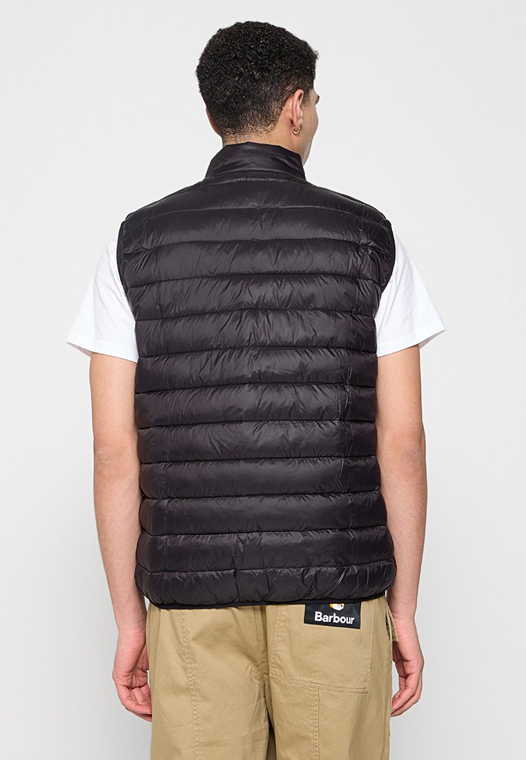 Schwarze, leichte Steppweste mit horizontalem gestepptem Design, hohem Kragen und ohne Ärmel. Über einem weißen T-Shirt getragen, mit minimalem Beschlag.