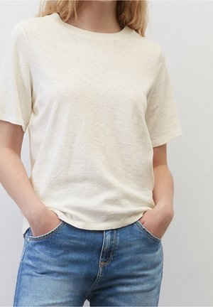 Vrouw draagt een effen crème-kleurig T-shirt met korte mouwen, lichtjes ingestopt in een hoog getailleerde blauwe jeans, met haar handen in de zakken.