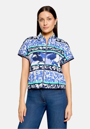 Vrouw met lang donker haar draagt een blauw patroon, korte mouwen, blouse met rits en blauwe jeans, staand tegen een effen lichte achtergrond.