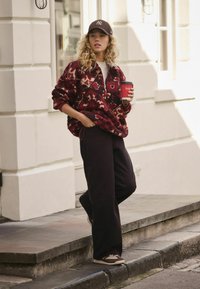 Fleece jacket in maroon with a floral pattern, paired with black wide-leg pants and brown sneakers. Holds a red coffee cup.