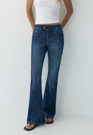 Femme portant un jean évasé bleu foncé, un haut blanc sans manches et des tongs noires, debout devant un fond clair uni.