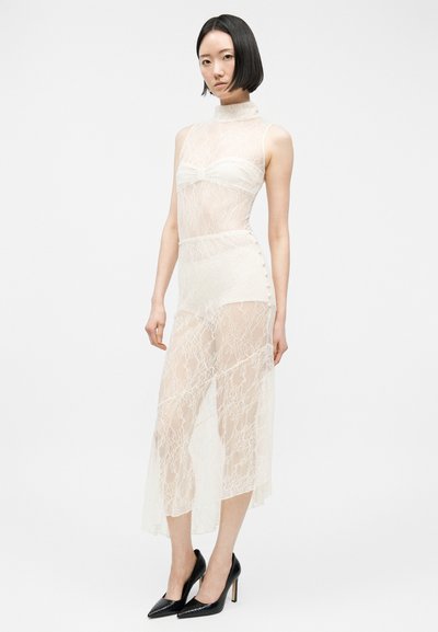 Femme aux cheveux courts noirs portant une robe blanche en dentelle transparente sans manches sur des sous-vêtements blancs et des escarpins noirs pointus, debout.