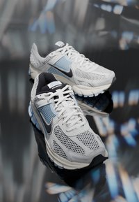Un par de zapatillas Nike Vomero 5 en blanco y azul claro con logotipos Swoosh negros, exhibidas sobre una superficie reflectante con un fondo desenfocado.