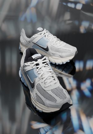 Une paire de chaussures de sport Nike Vomero 5 blanches et bleu clair avec des logos swoosh noirs, présentée sur une surface réfléchissante avec un arrière-plan flou.