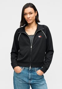 Μαύρο μπουφάν με φερμουάρ, από λείο ύφασμα με λευκές λεπτομέρειες, με μικρό λογότυπο στο στήθος, φορεμένο πάνω από γκρι μπλουζάκι και μπλε τζιν.