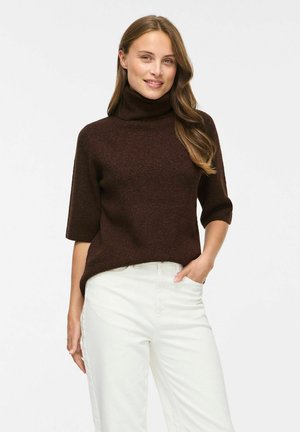 Femme portant un pull col roulé marron foncé avec des manches aux coudes et un pantalon blanc taille haute, debout avec une main dans la poche.