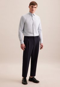 Camicia azzurra chiara con colletto a punta, abbinata a pantaloni eleganti a pieghe blu navy e scarpe nere stringate, su uno sfondo neutro.