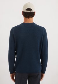 Marineblauwe gebreide trui met een textuurpatroon, lange mouwen en een ronde halslijn. Draagt een lichtgekleurde pet.