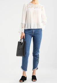 Blouse blanche avec accents en dentelle, jean à ourlet à franges bleu avec broderie verte, sac à main noir et mules noires à bout ouvert.