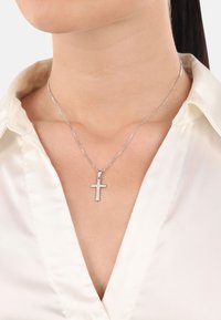 Silberner Kreuzanhänger an einer zarten Kette, getragen über einer satinierten weißen Bluse, die Einfachheit und Eleganz im Design hervorhebt.