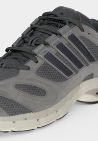 Γκρι αθλητικό παπούτσι Adidas με επάνω μέρος από διχτυωτό και σουέντ, μαύρες ρίγες, κορδόνια και μαλακή λευκή σόλα, απεικονισμένο πλάγια.