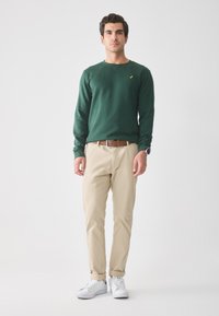 Uomo in piedi che indossa una camicia verde a maniche lunghe, pantaloni beige con una cintura marrone, sneakers bianche e un orologio da polso su uno sfondo semplice.