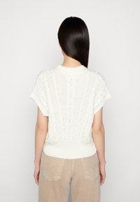 Weißer, kurzärmeliger Strickpullover mit strukturierten Zopfmustern; gerippter Saum und runder Ausschnitt; getragen mit beigen, hoch geschnittenen Hosen.