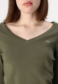 Olivengrønn v-hals genser laget av myk bomull, med en liten Levi's-lapp på venstre brystområde og en glatt tekstur med en tettsittende fasong.