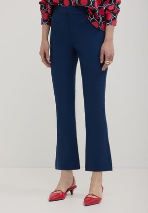 Eine Frau trägt marineblaue 3/4-Hosen kombiniert mit roten Kitten-Heels mit Slingback, goldenen Ringen und einer gemusterten roten und marineblauen Bluse.