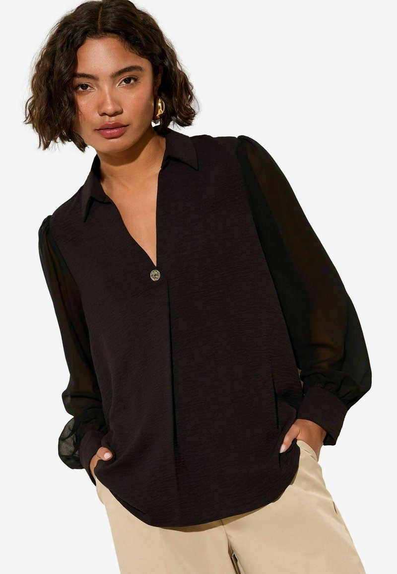 Blouse noire à manches longues avec un corps texturé et des manches transparentes, présentant un décolleté en V profond et un détail de bouton unique au col.