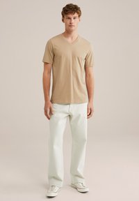 Svetlo rjavo v-neck majica v kombinaciji s svetlo zelenimi kavbojkami in belimi športnimi copati, z ožjim krojem in mehko teksturo. Preprost, sodoben stil.