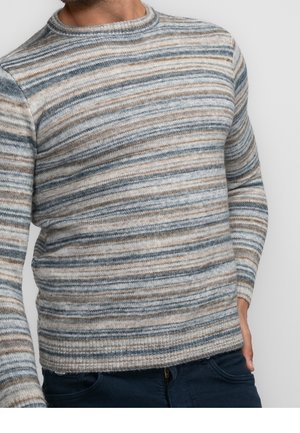 Mężczyzna w długim swetrze z dzianiny z poziomymi paskami w odcieniach szarości, beżu i jasnoniebieskiego, w parze z ciemnymi spodniami.