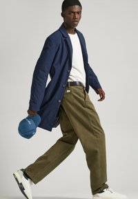 Jūras spēku garais mēteli, balta T-kreklā, olīvu zaļās biksēs, turētā zilu cepuri, ģērbs baltos un zaļos sporta apavos, staigājot neitrālā fonā.