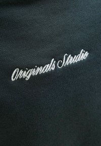 Fekete, sima textúrájú anyag, fehér, írott betűkkel hímzett "Originals Studio" felirattal. A dizájnelem közeli képe.