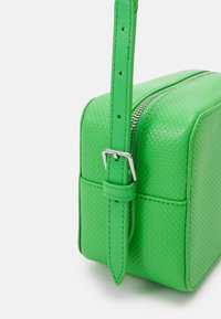Grüne strukturierte Handtasche mit rechteckiger Form, silberner Reißverschlussöffnung und verstellbarem Tragegurt mit einer Metallschnalle.
