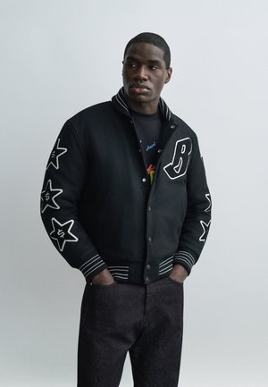 Joven con chaqueta universitaria negra con parches de estrellas blancas en las mangas y la letra "B" en el pecho, de pie con las manos en los bolsillos.