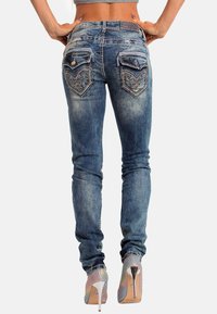 Jeans in denim blu aderenti con tasche posteriori decorative, leggere sfumature e dettagli di cucitura. Indossati con scarpe con tacco alto iridescenti.