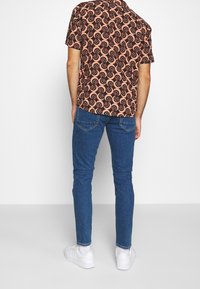 Camisa de manga corta para hombre con un patrón en espiral negro y rojo sobre un fondo melocotón, combinada con jeans azul ajustados y zapatillas blancas.