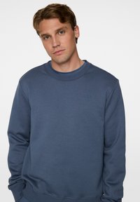 Sininen puuvillaneule, jossa on pyöreä kaula-aukko, ribbireunat hihoissa ja helmassa. Rinnassa on hillitty logo. Pehmeä pinta, rento muotoilu.