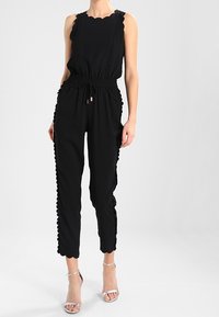 Combinaison noire à bords festonnés, sans manches, avec une taille élastique et des jambes fuselées. Portée avec des sandales à talons transparents.