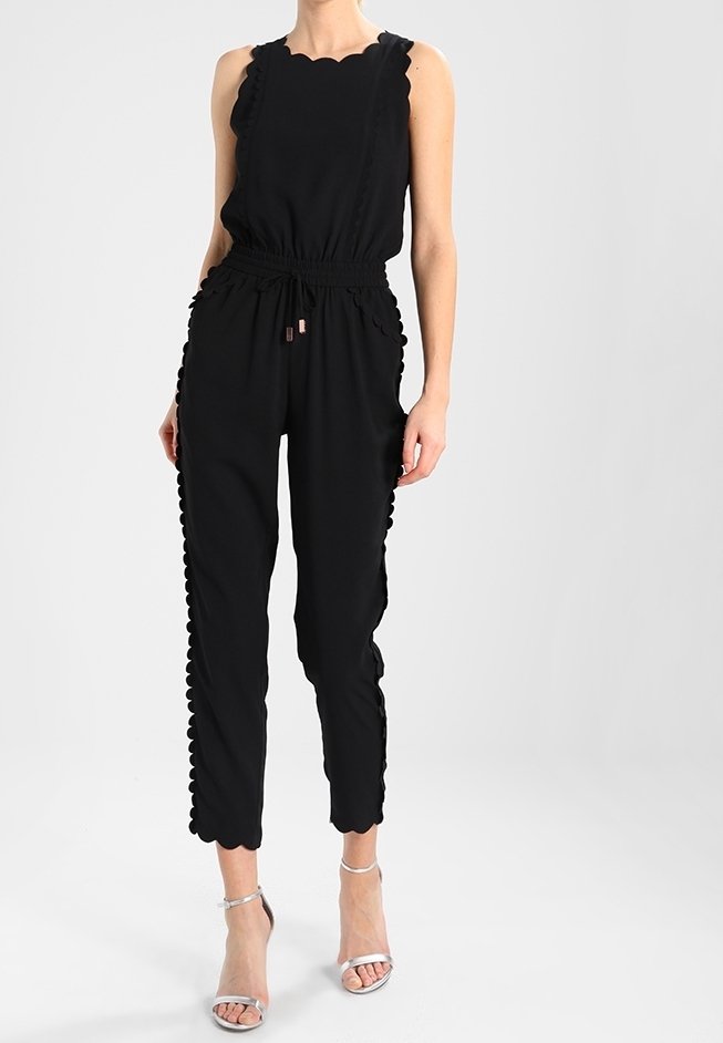 Combinaison noire à bords festonnés, sans manches, avec une taille élastique et des jambes fuselées. Portée avec des sandales à talons transparents.