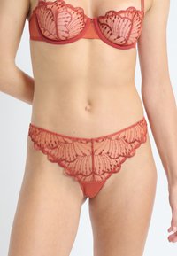 Soutien-gorge en dentelle rouge et culotte en dentelle assortie avec des bords festonnés, portés par une personne debout, montrés de la moitié du torse jusqu'aux cuisses supérieures.