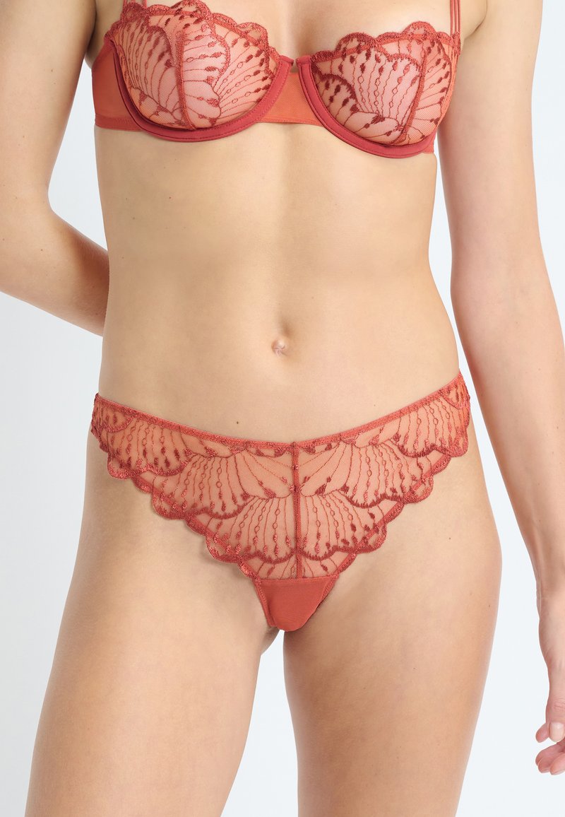 Roter Spitzenslip-BH und passende Spitzenslips mit gewellten Kanten, getragen von einer stehenden Person, die von der Mitte des Oberkörpers bis zu den oberen Oberschenkeln zu sehen ist.