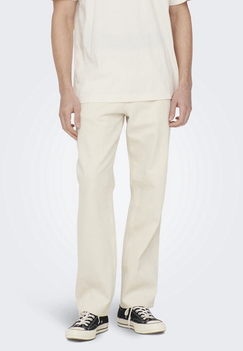 Pantalons droits couleur crème en coton, avec une texture lisse et des poches avant, associés à des baskets en toile noires.