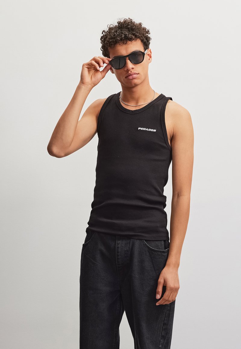 Giovane persona con capelli corti e ricci, che indossa occhiali da sole, un top nero senza maniche con la scritta ""PEGADOR"" e pantaloni scuri, in piedi su uno sfondo neutro.