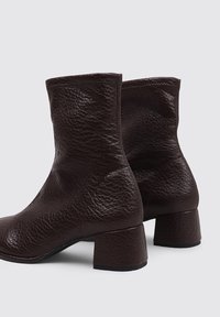 Botas de cuero marrón hasta el tobillo con un acabado texturizado, que cuentan con un tacón cuadrado y un ajuste ceñido alrededor del tobillo. Diseño minimalista sin adornos.