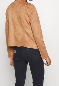 Persona che indossa una giacca in suede beige e jeans attillati blu scuro, vista da dietro su uno sfondo chiaro e semplice.