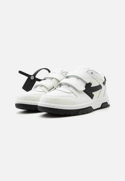 Λευκά αθλητικά παπούτσια χαμηλού ύψους με μαύρο σχέδιο βέλους, δύο λουριά με βέλκρο, χοντρή μαύρη σόλα και προσαρτημένη μαύρη ετικέτα στο αριστερό παπούτσι.