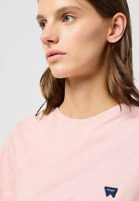 T-shirt di cotone rosa con scollatura rotonda e maniche corte, dotata di una piccola etichetta logata Wrangler blu nell'area del petto sinistro.