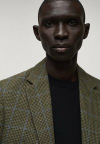 Blazer a quadri verdi con dettagli blu, rever strutturati e una finitura texturizzata, indossato sopra un maglione nero. Design pulito e moderno.