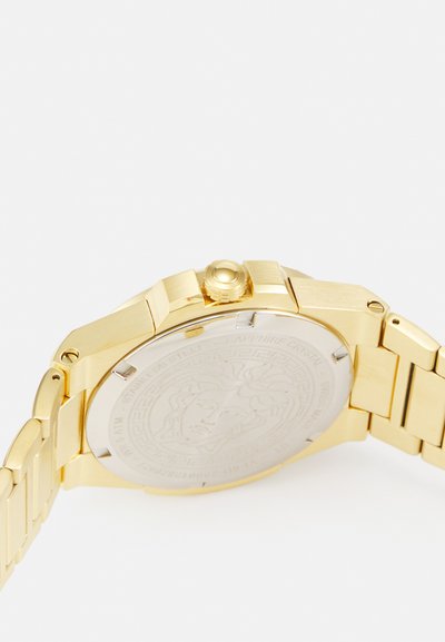 Zelta tonis pulkstenis ar nerūsējošā tērauda korpusu, aizmugurē iegravētu dizainu un saskaņotu metāla ķēdes aproci vienotam izskatam.