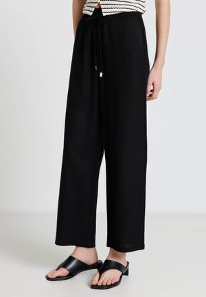 Frau trägt lockere schwarze Hosen mit Kordelzug und schwarze Sandalen mit Absatz, steht auf einem hellgrauen Boden vor einem schlichten weißen Hintergrund.