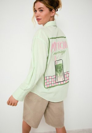 Jeune femme aux cheveux blonds portant une chemise rayée vert clair avec l'inscription « Matcha mornings » et un motif de verre au dos, assortie à un short beige.