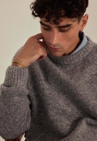 Pull gris texturé en tricot avec un ourlet côtelé, porté avec une chemise à col bleu clair en dessous. Gros plan sur une personne reposant son menton sur sa main.