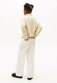 Beiger, kurzer Pullover, weiße weite Hose und schwarze Schuhe. Das Outfit zeichnet sich durch einen geschichteten Look mit einem kragenlosen Hemd darunter aus.