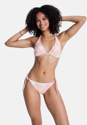 Junge Frau mit lockigem Haar lächelt, trägt einen pink-grünen Blumenbikini und posiert mit den Händen hinter dem Kopf vor einem einfarbigen Hintergrund.